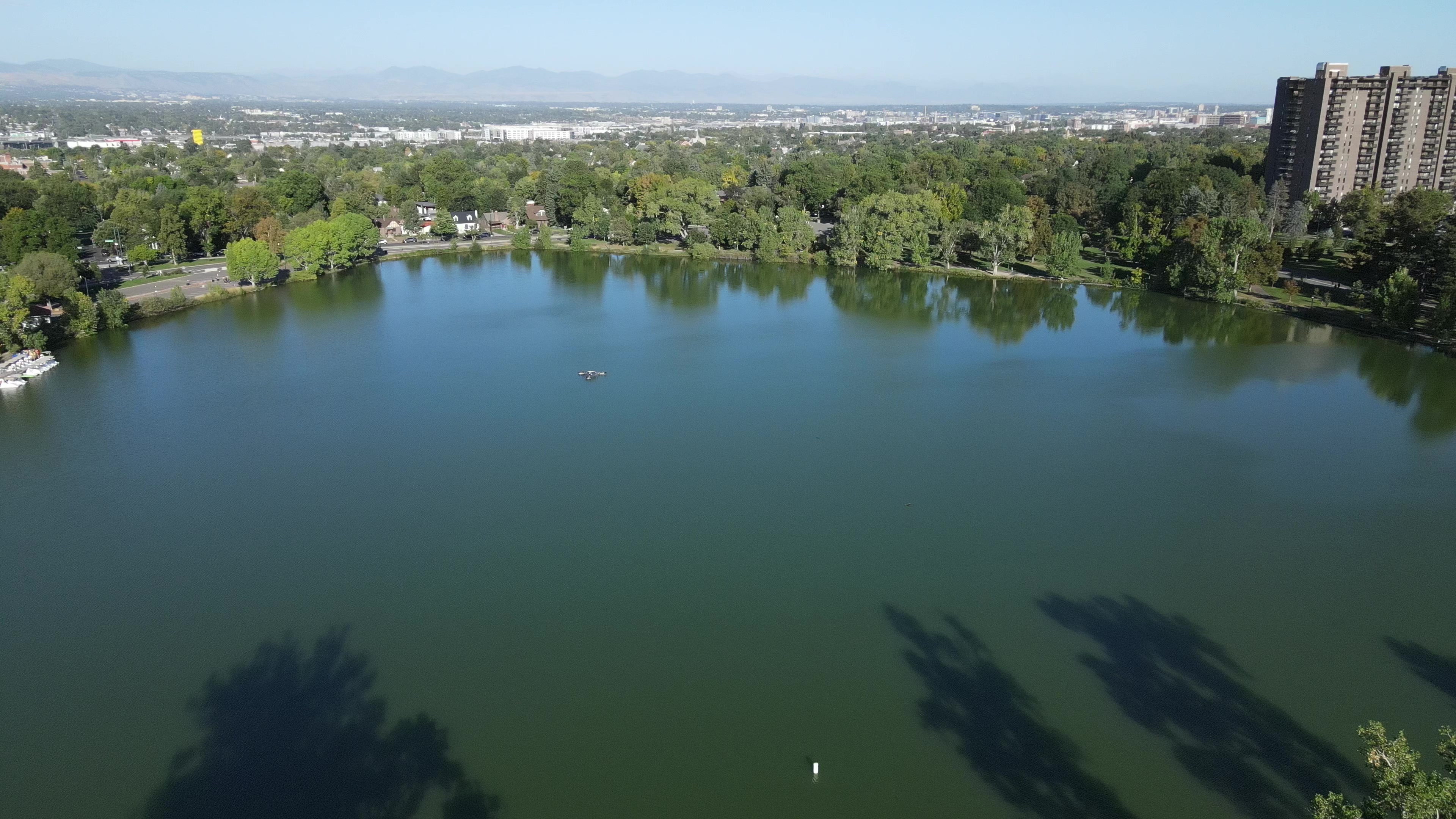 Aerial view over Smith Lake in Washington Park, Denver: a lone paddler on calm water with the downtown skyline and Rocky Mountain Front Range on the horizon under a clear blue sky.